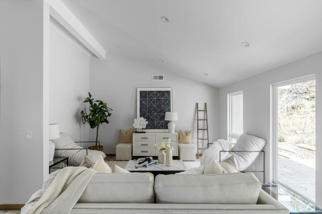 A bright white small living room with neutral furniture, minimal decor, and large windows flooding the space with light.